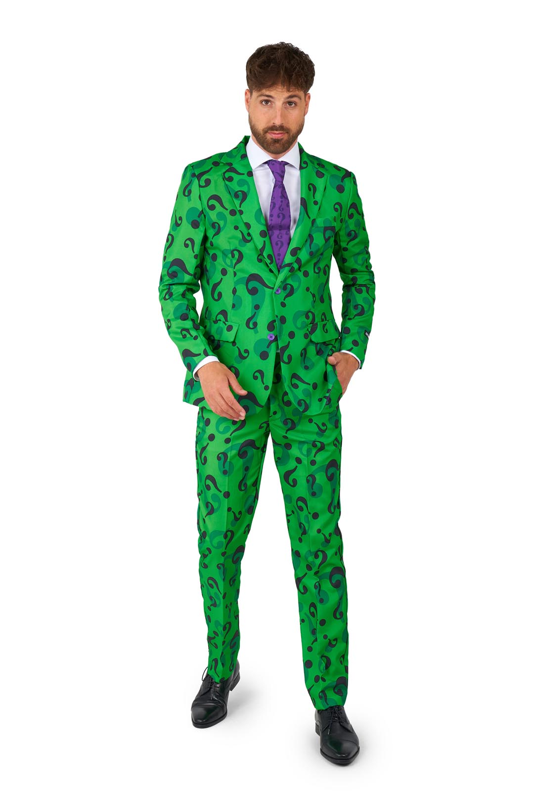 A man dressed in The Riddler suit by Suitmeister, complete with a bright green jacket adorned with question marks, a white shirt, and a purple tie, stands on a plain white background with one hand in his pocket.