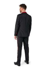 A man is facing away from the camera, dressed in a Suitmeister Harry Potter Suit Gryffindor™, featuring black fabric adorned with red trim details. He sports short hair and pairs his outfit with black shoes. The background is a simple white setting.