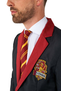 A man dressed in a Suitmeister Harry Potter Gryffindor™ Suit, featuring a dark blazer adorned with a red lapel and the Gryffindor crest. He completes his look with a white shirt and a red and yellow striped tie, all set against a plain white background.