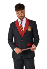 A man dressed in the Harry Potter Gryffindor™ Suit by Suitmeister, featuring a black design with red accents and a striped tie. He's holding a wand and wearing round glasses, with a fantasy-themed crest patch on the blazer.