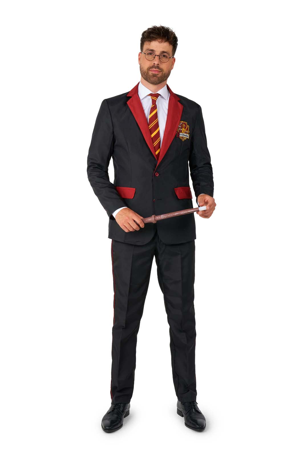 A man stands wearing the Suitmeister Harry Potter Gryffindor™ Suit, featuring maroon accents, a maroon and yellow striped tie, and glasses. He holds a wand in both hands, with an embroidered crest on the pocket of his black suit.