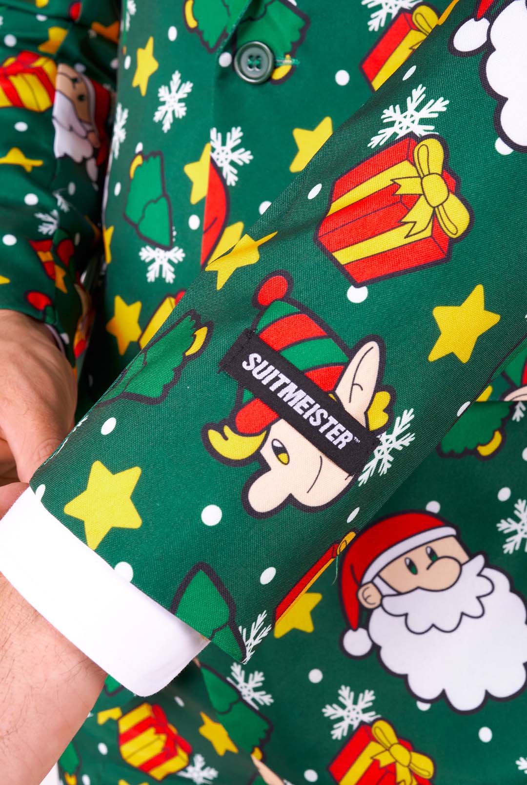 Close-up of a person's arm wearing the Santa Elves Green suit from Suitmeister, featuring a festive green design adorned with colorful Christmas-themed prints like Santa faces, gifts, stars, and Christmas trees. The sleeve is labeled with the Suitmeister brand.