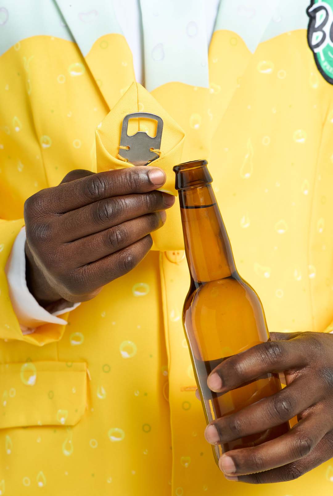 A person wearing a Suitmeister Premium Beer Yellow suit, which features a design inspired by the beverage, is holding a brown beer bottle in one hand. With the other hand, they are pulling a bottle opener from a pocket on the suit. The background is plain white.