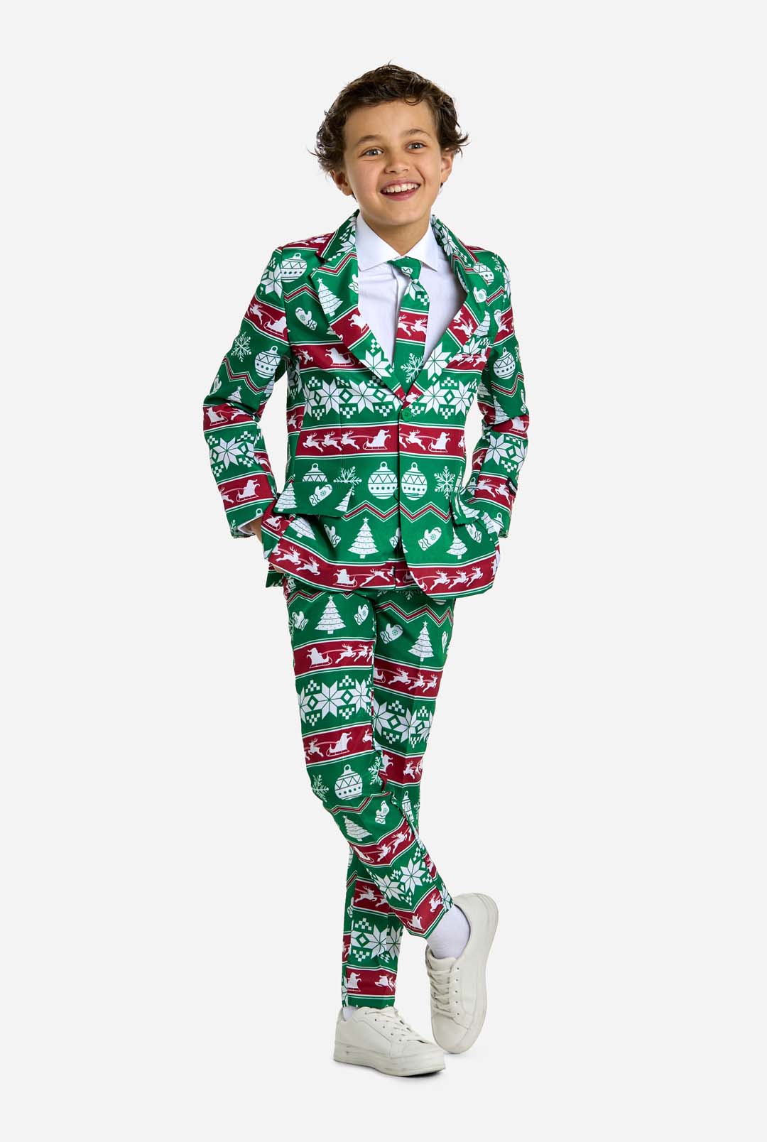 A child clad in the "Christmas Suit Green Nordic" by Suitmeister, featuring Christmas-themed patterns like trees and snowflakes, stands smiling with one leg crossed over the other. The suit showcases a predominantly green color with red and white accents, perfectly paired with white sneakers.