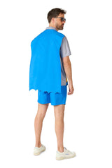 A man wearing sunglasses is styled in the Batman™ - summer set from Suitmeister, featuring a blue and grey ensemble that includes a shirt, shorts, and a cape. He stands looking to the side with white sneakers rounding out the look against a white background.