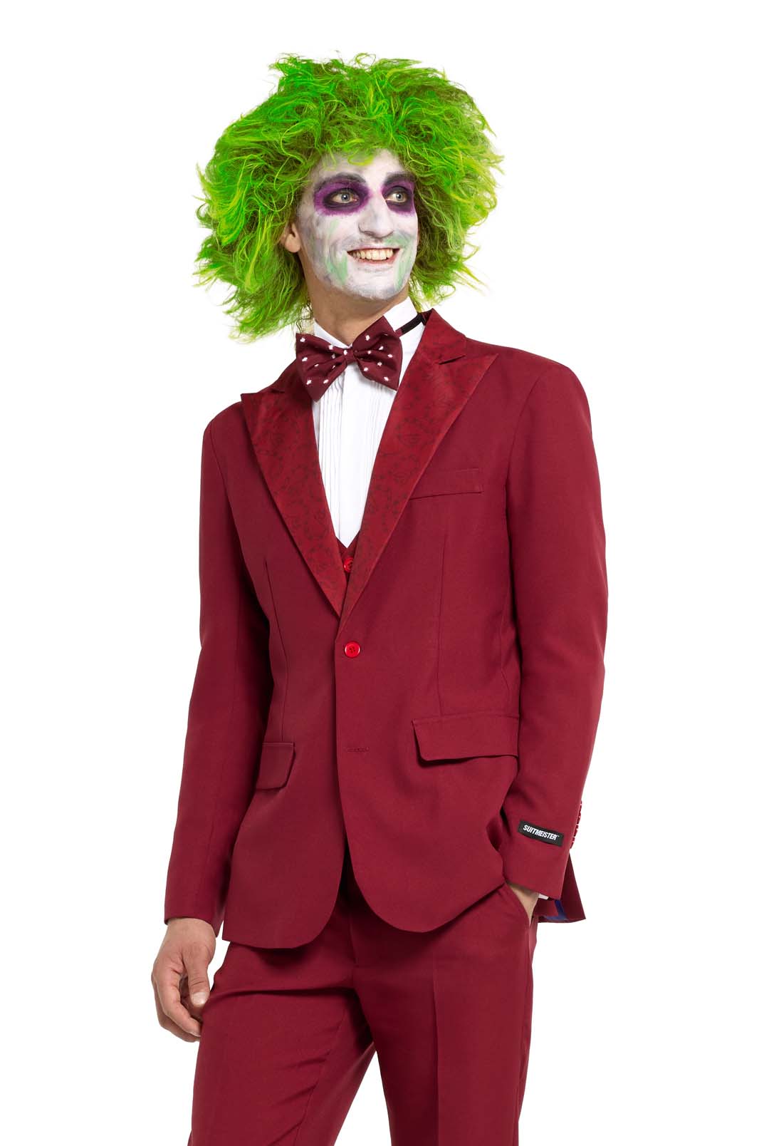 A person in a vivid green wig and white face paint with purple accents is wearing a Beetlejuice Groom costume from the brand Suitmeister, featuring a red suit, white shirt, and polka dot bow tie. They are smiling and standing against a white background.