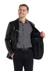 A person smiling while wearing a Suitmeister Sequin Blazer Black, paired with a gray button-up shirt and black pants. One hand is in their pocket as they hold the blazer open with the other against a plain white background.