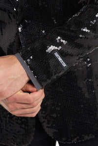 A close-up shot captures a person wearing the Suitmeister Sequin Blazer Black. The visible sleeve sports a gray label with the text STITCH EDITER. One hand is adjusting the cuff, showcasing the blazer's intricate sequin details.