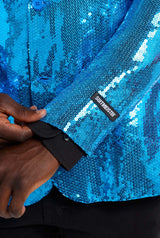 A person adjusts the cuff of a shimmering Suitmeister Sequins Blue Blazer. The cuff features a black label adorned with white text, providing a striking contrast to the person's dark wrist and black inner sleeve.