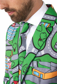 A man is dressed in a Suitmeister ensemble called "Carpet City Green," characterized by its vibrant design featuring a cartoon city map with roads, cars, buildings, and trees. The outfit includes a matching tie and is paired with a white shirt underneath. The emphasis is on the detailed pattern and overall style of the outfit.