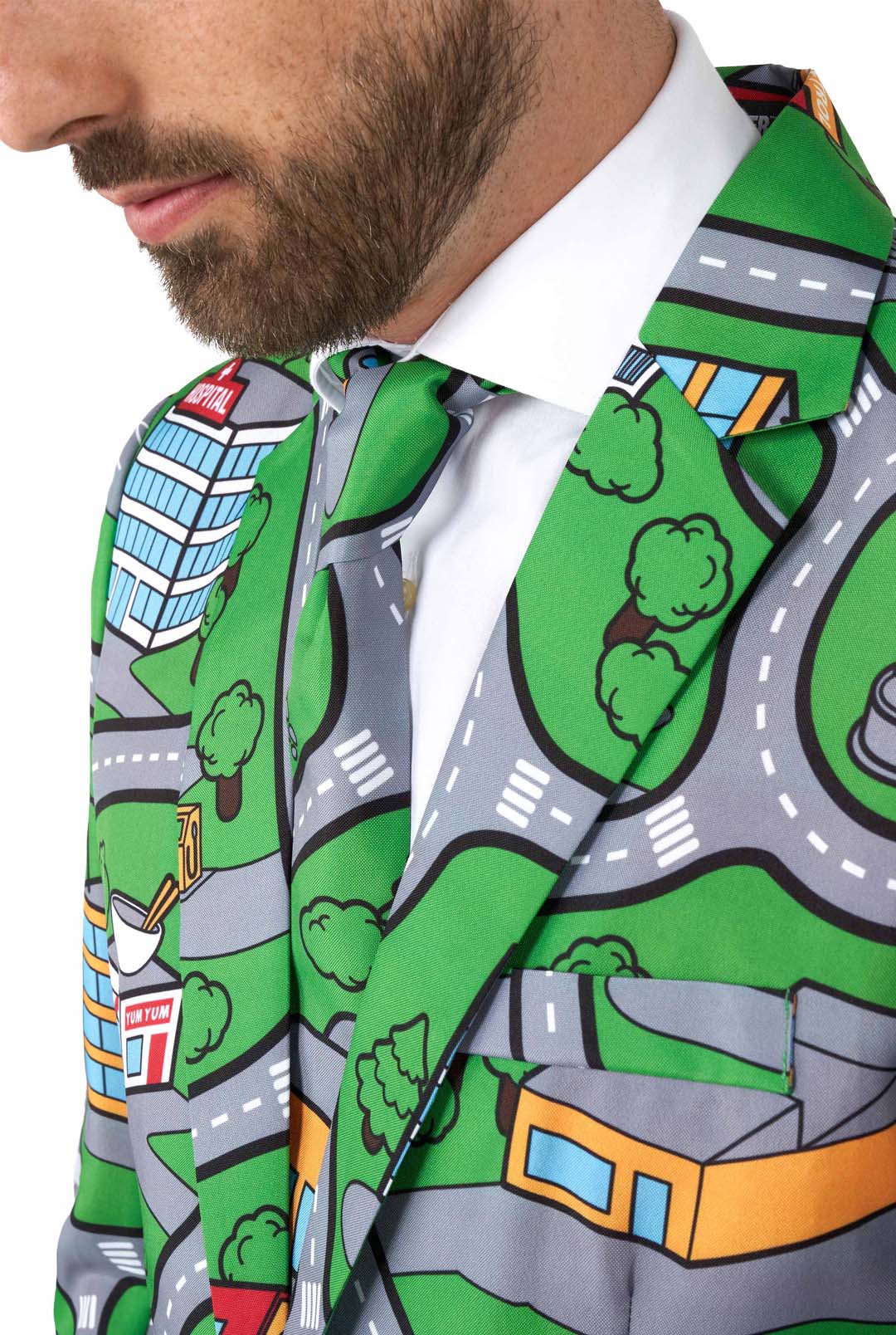 A man is dressed in a Suitmeister ensemble called "Carpet City Green," characterized by its vibrant design featuring a cartoon city map with roads, cars, buildings, and trees. The outfit includes a matching tie and is paired with a white shirt underneath. The emphasis is on the detailed pattern and overall style of the outfit.