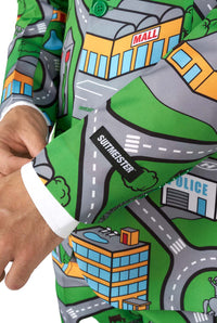 A person adjusts the cuff of a vibrant Suitmeister suit called Carpet City Green, featuring a cartoon-style city map design. The map includes buildings labeled Mall and Police, with roads winding throughout. The suit boasts a colorful and playful design with shades of green, gray, and blue.