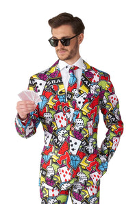 A man in dark sunglasses and a vibrant Casino Icons suit from Suitmeister, adorned with a casino-themed pattern featuring dice, playing cards, and slot machine symbols, holds a set of playing cards.