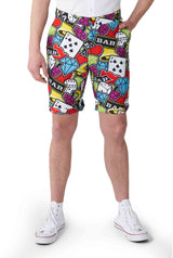 A person is sporting a vibrant pair of shorts from Suitmeister's Casino Icons - Carnival suit collection, featuring lively patterns of dice, cards, and bars. They have their hands tucked into the pockets and are paired with white sneakers against a plain white backdrop.