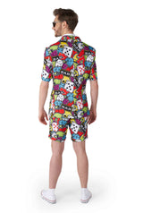 A man dressed in the vibrant "Casino Icons - Carnival Suit" by Suitmeister is adorned with patterns of playing cards, dice, cherries, and other symbolic designs. He stands facing away, wearing a short-sleeved top and matching shorts. His ensemble is completed with sunglasses and white shoes.