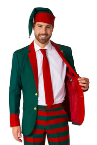 A man is dressed in the Suitmeister Santa's Elf Green - Christmas Elf Suit, featuring a festive pointy hat and striped pants. He is holding open the jacket to showcase a red lining inside.