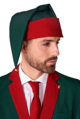 A man with a beard is dressed in Suitmeister's "Santa's Elf Green" ensemble, complete with a festive green suit adorned with red accents and a matching elf-like hat featuring a green hue and red band. He complements his look with a red tie while gazing off to the side.