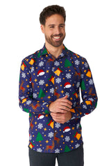 A smiling man is wearing the "Christmas Icons Blue" shirt by Suitmeister, featuring a festive array of Christmas-themed patterns such as snowflakes, reindeer, Santa hats, Christmas trees, and ornaments set against a dark background. He is clasping his hands in front.