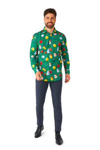 A person stands smiling, dressed in the "Christmas Ornaments Green" Christmas shirt by Suitmeister, featuring holiday-themed designs like snowmen and ornaments. They are also wearing dark blue pants and black shoes against a white backdrop.