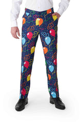 A person is wearing the Suitmeister Confetti Balloons Navy pants, featuring colorful balloon and streamer patterns on a dark blue background. They are also wearing a white shirt and black shoes.