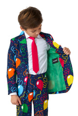 A young boy wearing the vibrant "Confetti Balloons Navy" suit from Suitmeister, which features lively balloon and confetti patterns, examines the inside of his jacket. The ensemble is complemented by a bright pink tie and a crisp white shirt, with the jacket's interior showcasing a bold green lining.