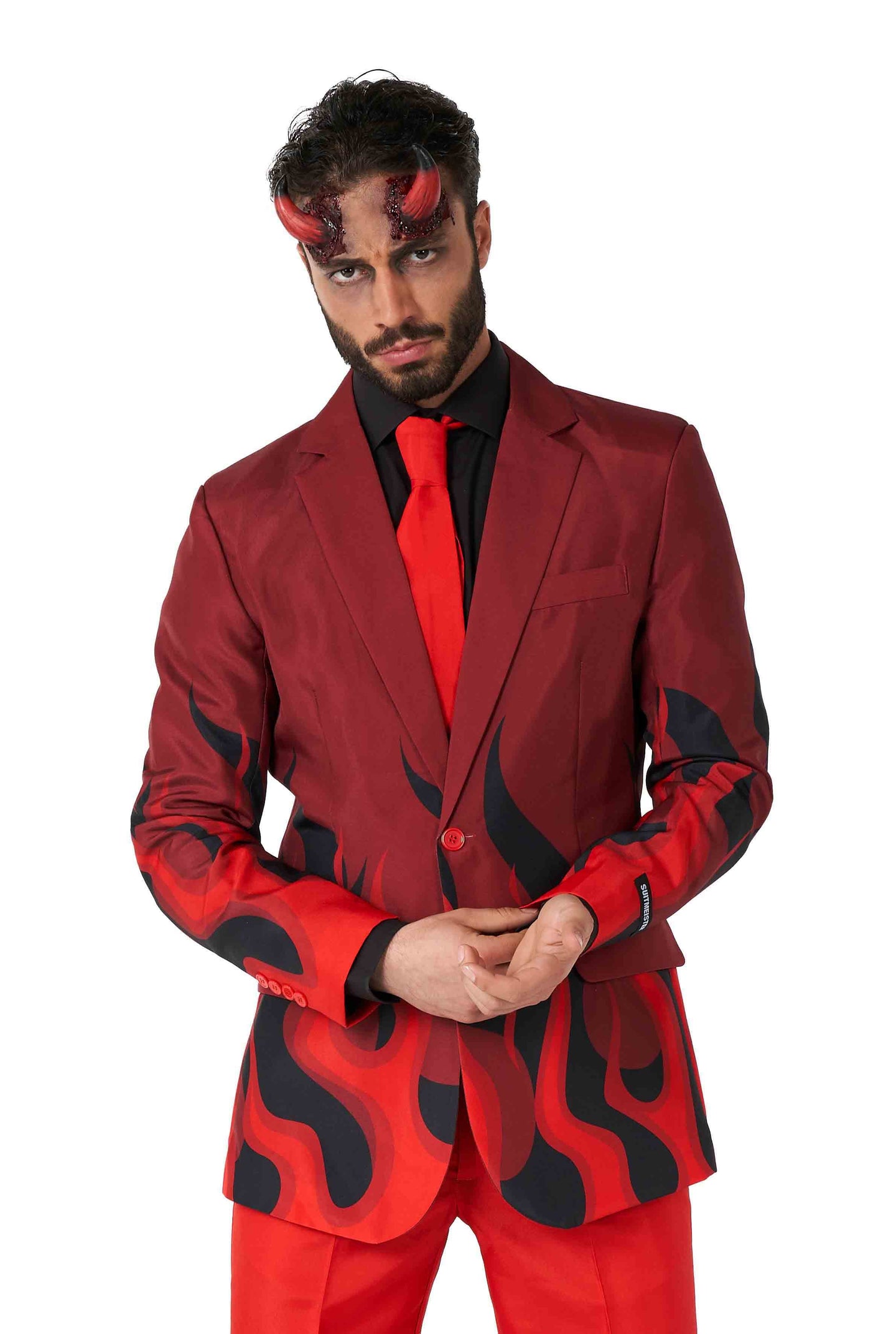 A man dressed in the Devil Red Halloween suit by Suitmeister poses against a white background. His ensemble includes a vibrant red tie and a sleek black shirt, topped off with small red devil horns, all while maintaining a serious expression. The suit itself features striking black flame patterns.