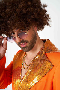 A person with curly hair and a beard, wearing the Suitmeister Disco Suit Orange adorned with shiny gold sequins, gazes over sunglasses with a playful expression against a plain white background.