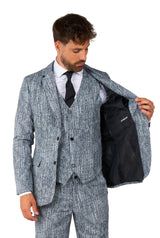 A man dressed in the Peaky Blinders costume "20's Gangster Grey" suit by Suitmeister, paired with a black tie and white shirt, opens his suit jacket to reveal the lining. He sports short hair and a beard against a plain white backdrop.