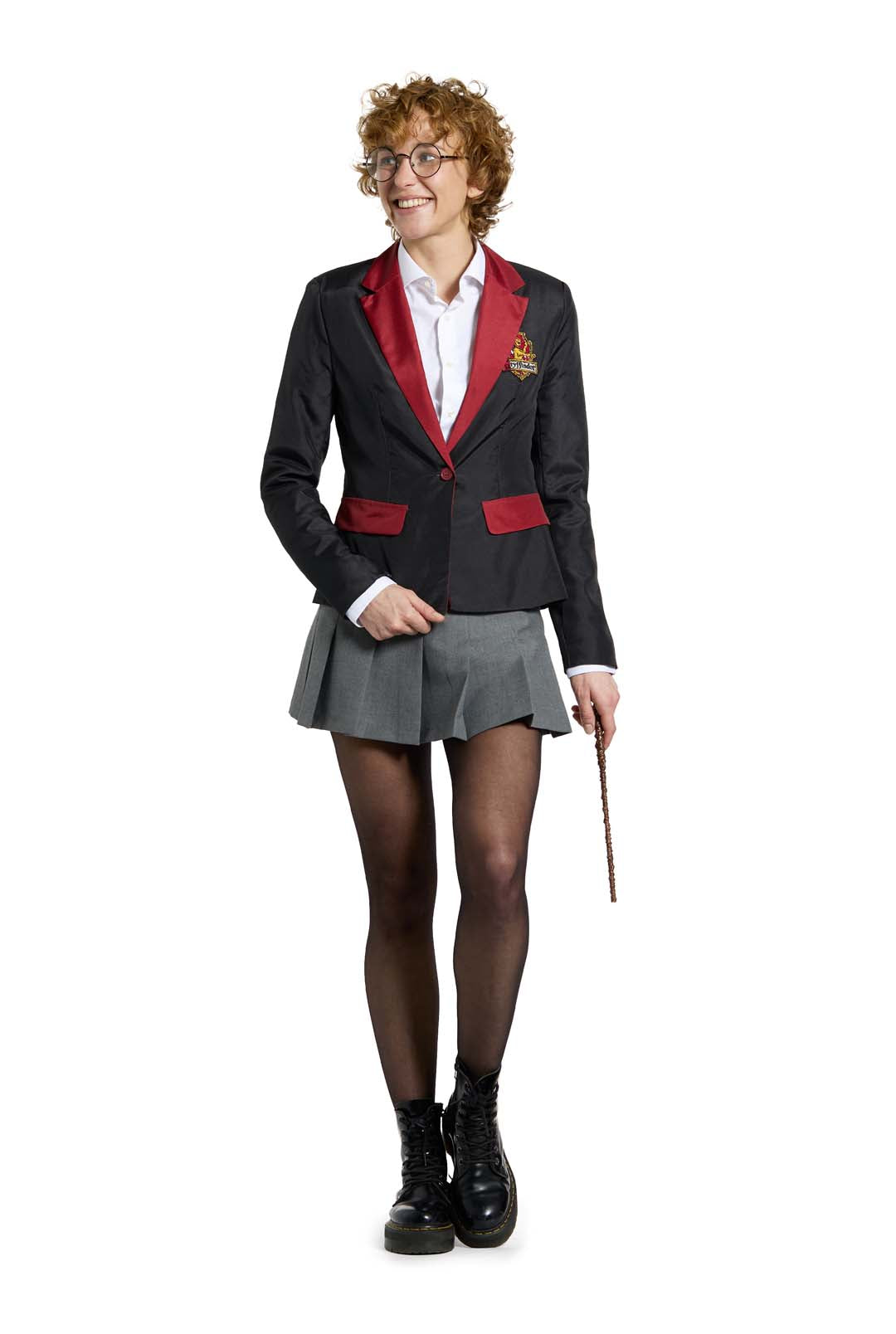 An individual with curly red hair and glasses is wearing the Suitmeister Harry Potter Gryffindor™ Blazer, paired with a white shirt, gray skirt, black stockings, and boots, holding a wand in their right hand on a plain white background.