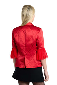 A person with long blonde hair is wearing the Harley Quinn™ red satin jacket by Suitmeister, showcasing a diamond pattern and sheer ruffled sleeves. They have paired their outfit with a black skirt and sheer black stockings, standing with their back to the camera.