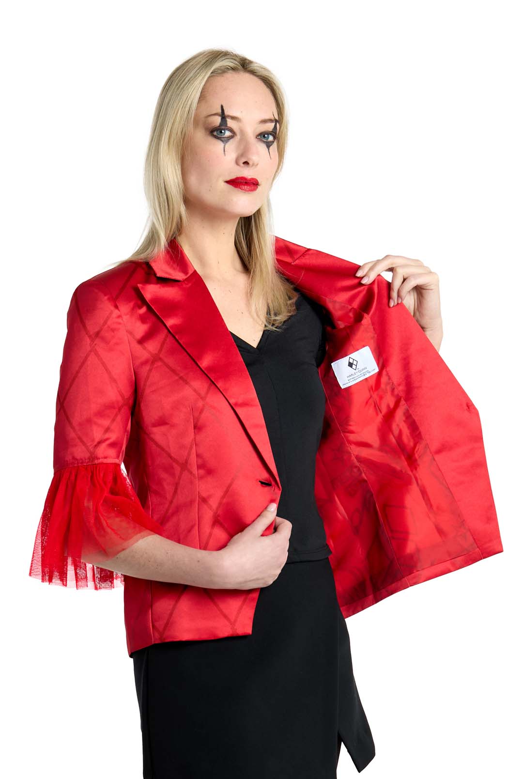 A woman with dramatic eye makeup is wearing the Harley Quinn™ jacket by Suitmeister, featuring quilted and lace details. She holds the jacket open to reveal the label inside. Complementing her look, she sports a black top and skirt while posing against a white background.