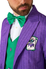 A person wearing the Joker™ Tailcoat Suit Purple from Suitmeister, paired with a green bow tie and vest. The pocket of the suit showcases an embroidered patch of a cartoonish joker figure.