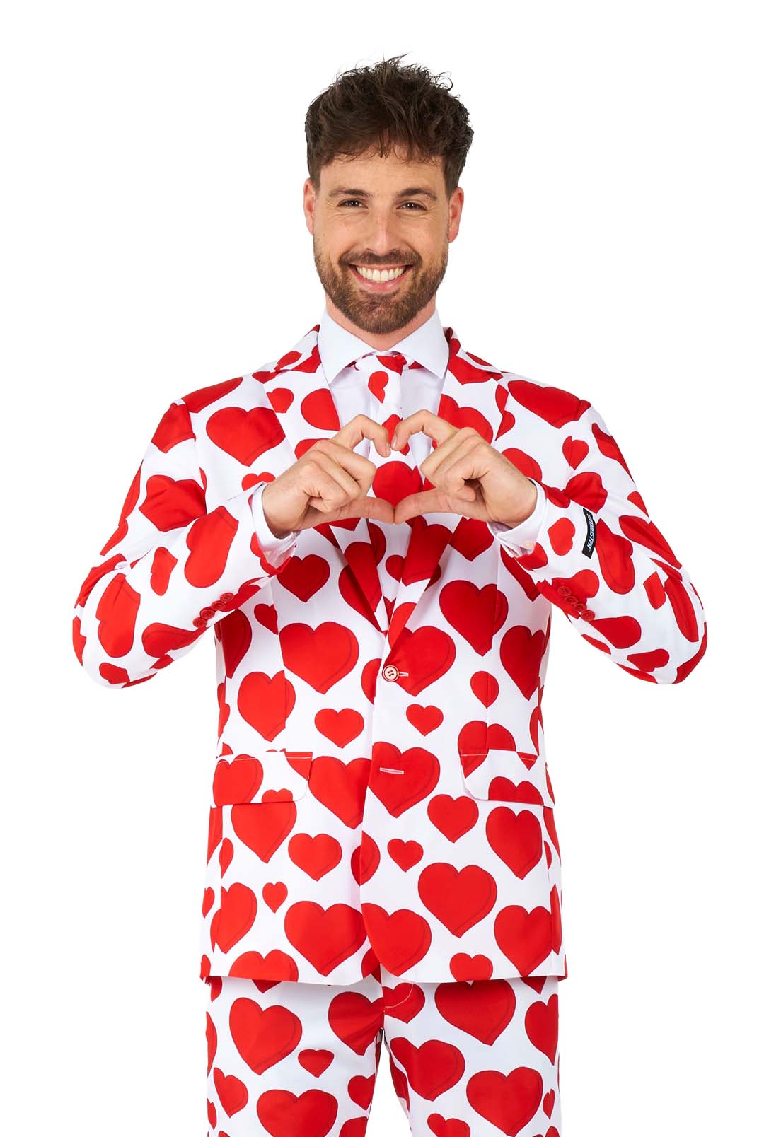 A man smiling and wearing a Suitmeister Love - Heart suit, which is a white suit adorned with red heart patterns. He is forming a heart shape with his hands in front of his chest against a plain white background.