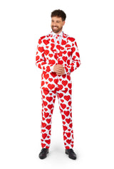 A man stands smiling, wearing the Suitmeister's "Love - Heart Suit" featuring a white suit with red heart patterns. He has curly hair and a beard, complemented by a white shirt and a matching patterned tie, all set against a plain white background.