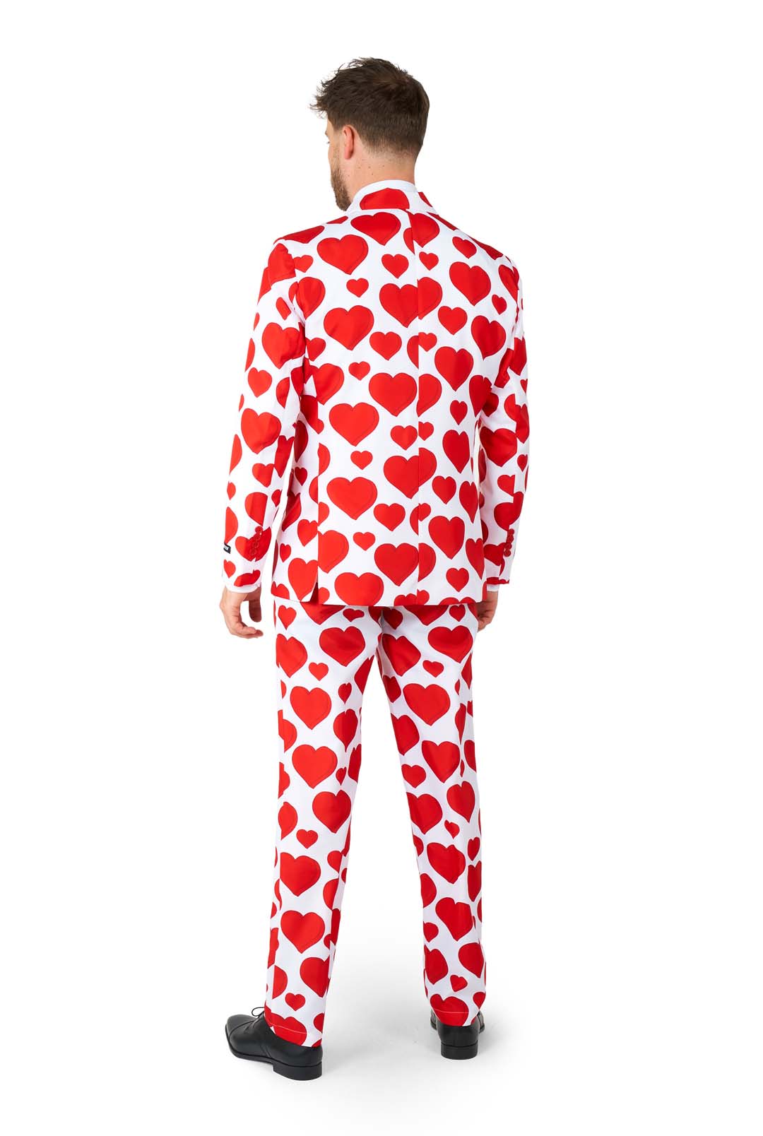 A man is standing facing away wearing the "Love - Heart suit" by Suitmeister. The predominantly white suit features bold red heart patterns. He has paired it with black shoes, and the background is plain white.