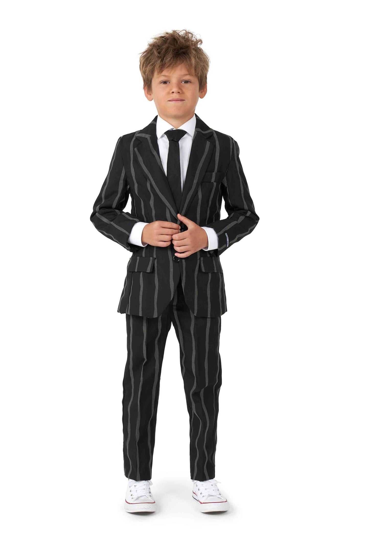 A young boy with messy hair stands confidently in an oversized pinstripe black suit from Suitmeister, paired with a white shirt and black tie. He is wearing white sneakers, smiling, and posing against a plain white background.