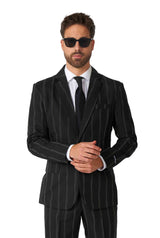 A man wearing the "Oversized Pinstripe Black" suit by Suitmeister stands confidently against a plain white background. He has sunglasses on, a well-groomed beard, and is adjusting his suit jacket with his hands.