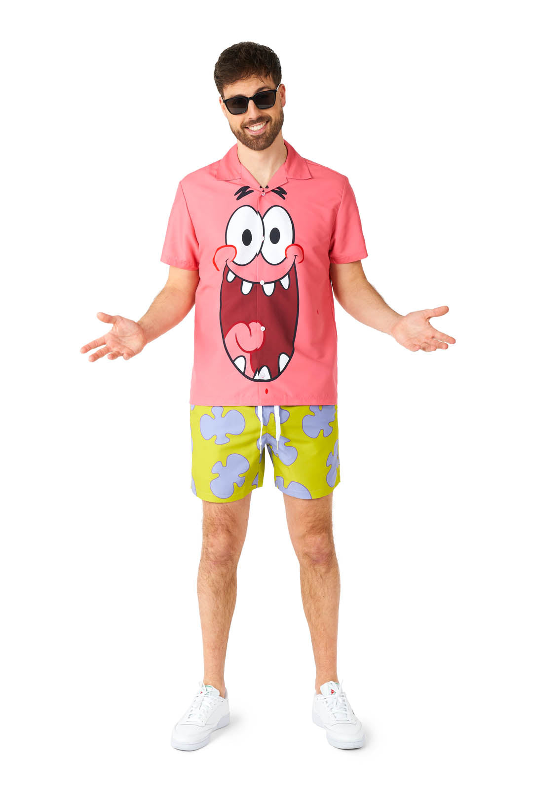 A man dressed in the Suitmeister Patrick costume, featuring sunglasses, a pink shirt with a cartoon face design, and green shorts with cloud patterns, poses with his arms slightly outstretched. He is wearing white sneakers and stands against a plain white background.