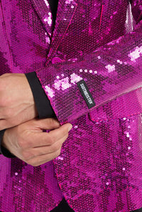 A close-up shot showcases an individual wearing a Sequins Blazer Pink by Suitmeister, with their left hand adjusting the shimmering sleeve. A black tag on the sleeve displays the brand name, Suitmeister. Underneath, they are dressed in a black shirt.