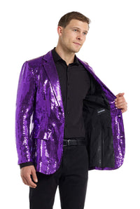 A man is dressed in a vibrant, shiny purple Sequins Blazer by Suitmeister, layered over a black shirt and pants. He holds one side of the blazer open, revealing its interior lining against a plain white background.