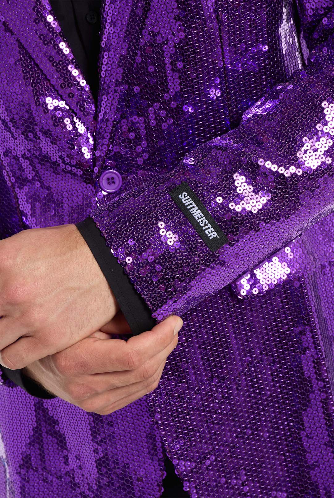 A person wearing a vibrant Sequins Blazer Purple by Suitmeister adjusts the sleeve. A black tag featuring the white text "SULTNT WESTER" is visible on the cuff, standing out against the plain background and highlighting the blazer's sparkling purple sequined texture.