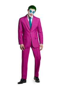 A person dressed in the Ruby Red Joker suit by Suitmeister, complete with a vibrant pink jacket and trousers, paired with a turquoise tie and black shoes, is posing against a white background. Their face is painted with blue eye shapes and red lips, complemented by their green hair.