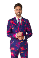 A man grins while sporting the Suitmeister brand's bold Retro Neon Navy - Carnival suit, showcasing a vibrant pattern of flamingos and palm trees on a dark purple background. He stands against a simple white backdrop, adjusting his left sleeve.