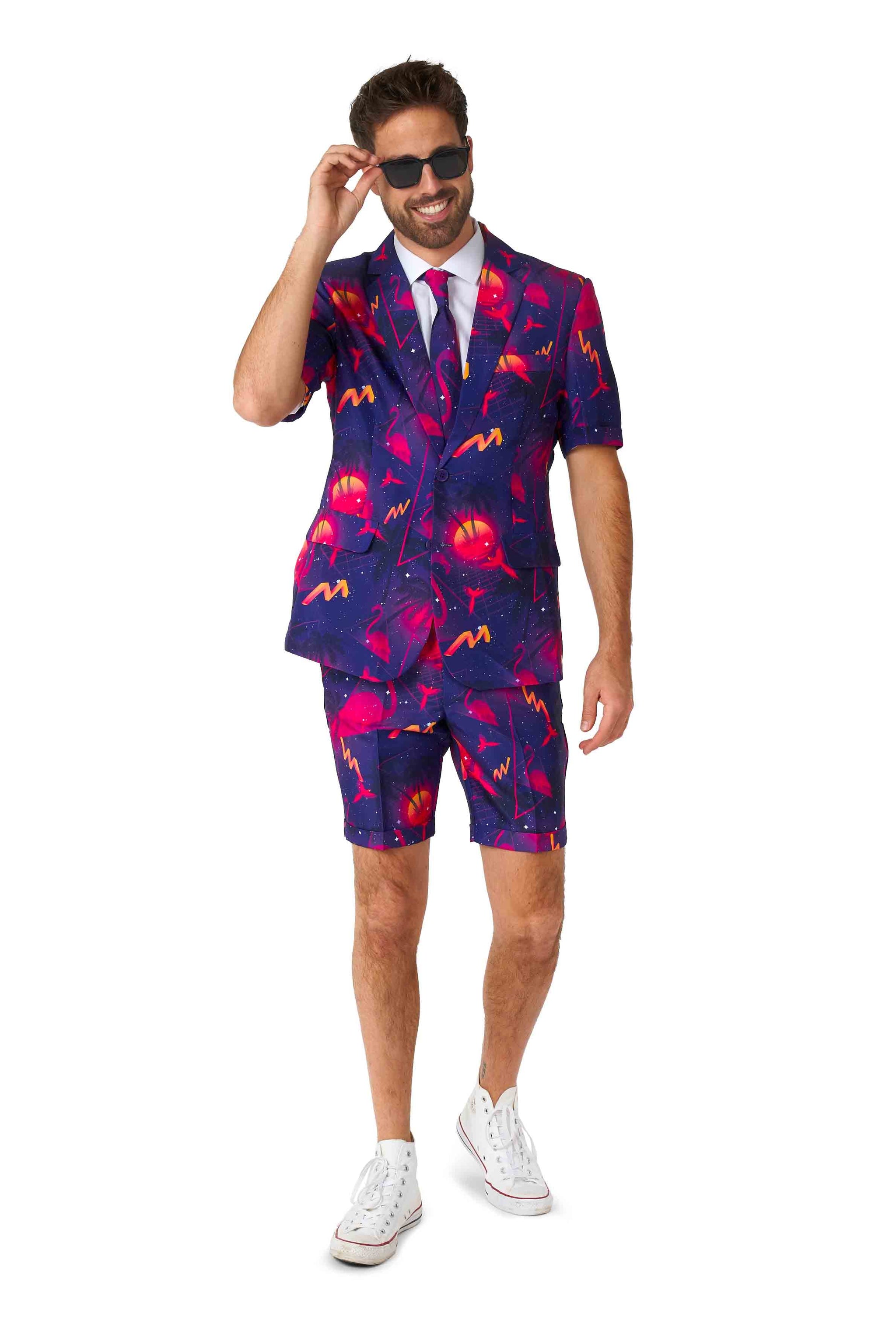 A man wearing the Suitmeister Retro Neon Navy - Carnival suit, featuring a vibrant and colorful design with shorts in neon pink and blue. He sports sunglasses, a white shirt, and white sneakers, smiling confidently as he adjusts his eyewear.