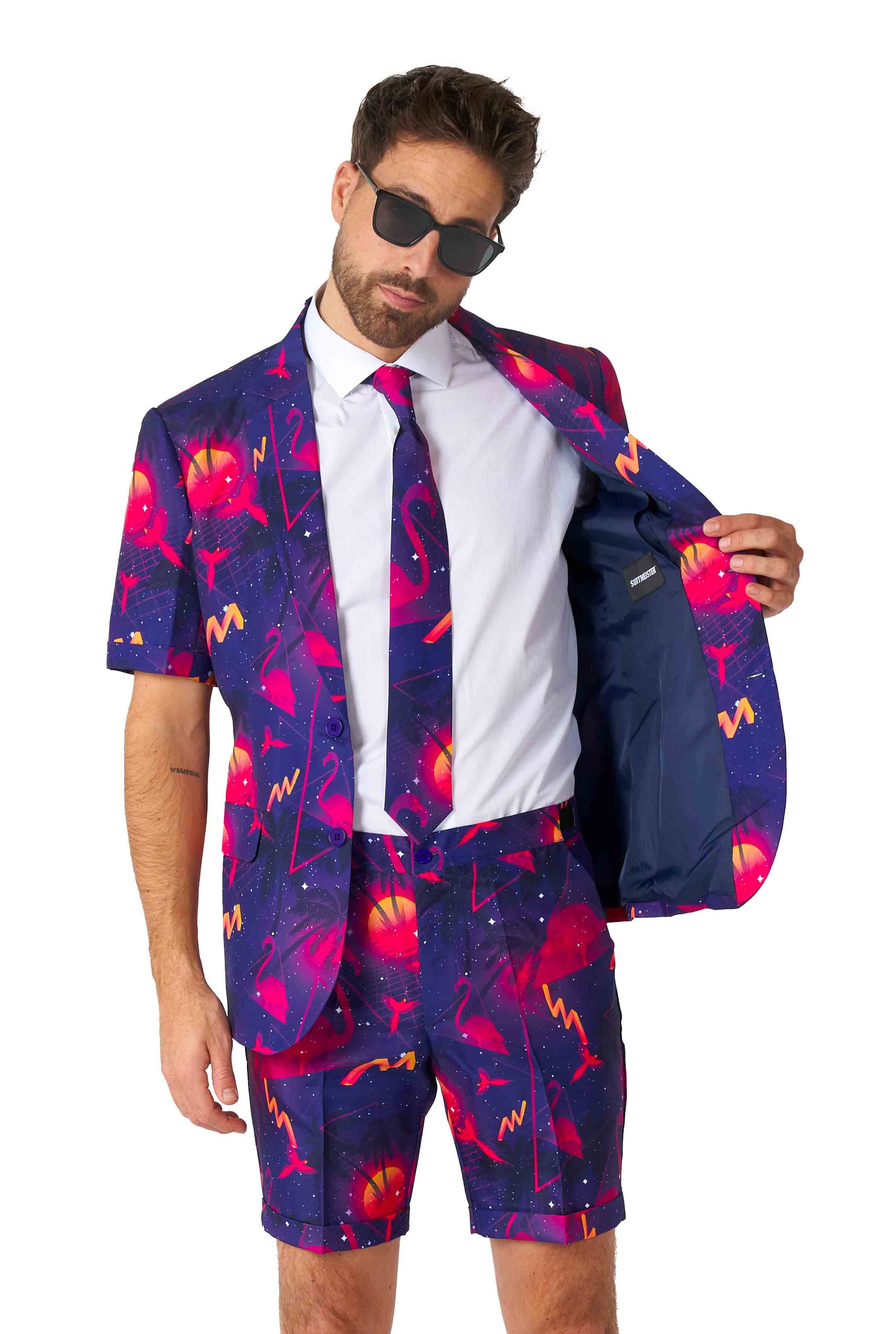 A man wearing sunglasses is dressed in the Retro Neon Navy - Carnival suit by Suitmeister, featuring a galaxy pattern with short sleeves. The suit also includes matching shorts and a tie, and he reveals the inner lining of his open jacket.