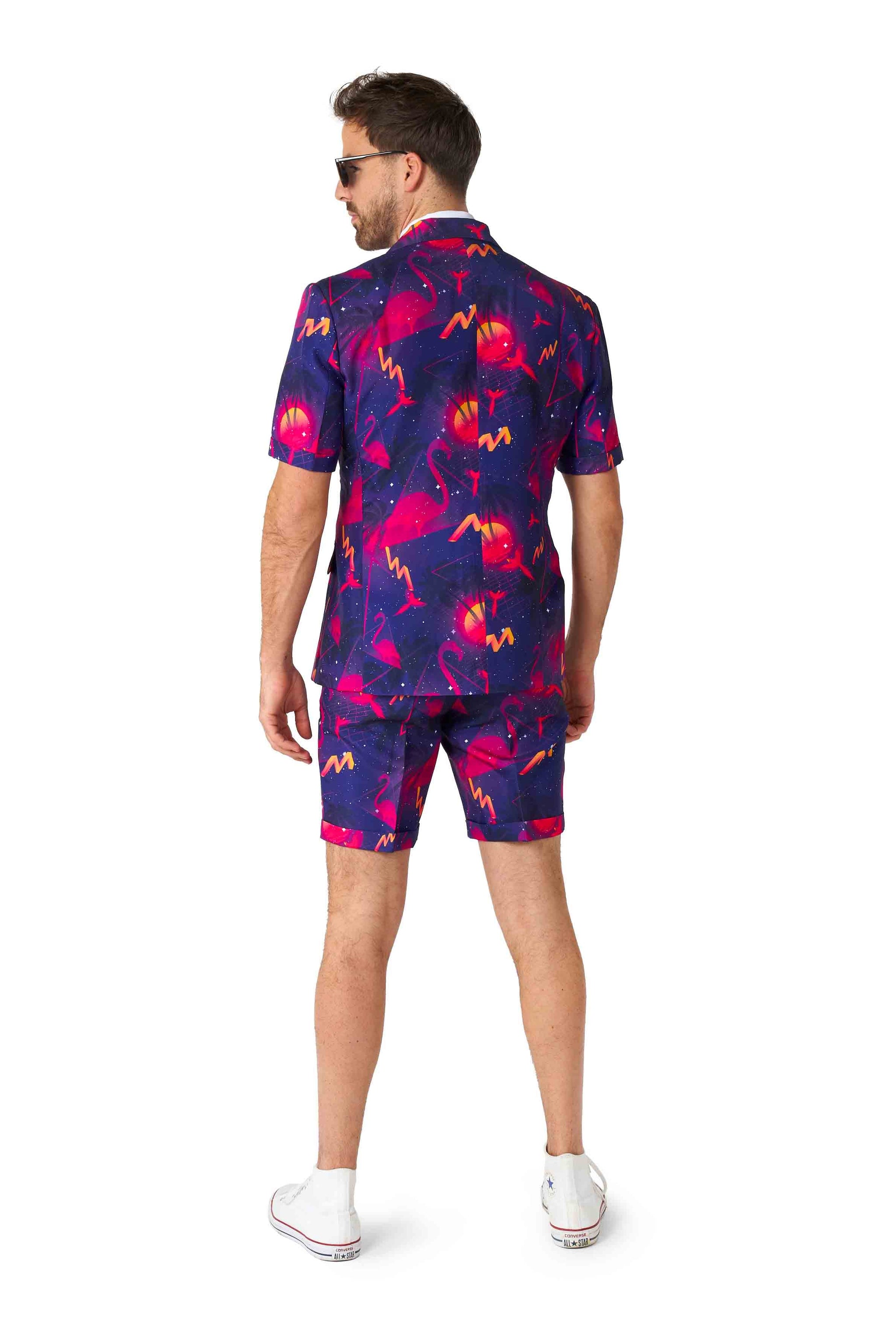 A person wearing the vibrant Retro Neon Navy - Carnival suit by Suitmeister, featuring a tropical pattern of pink flamingos and palm trees against a dark background, stands facing away. They are also sporting white sneakers and sunglasses.