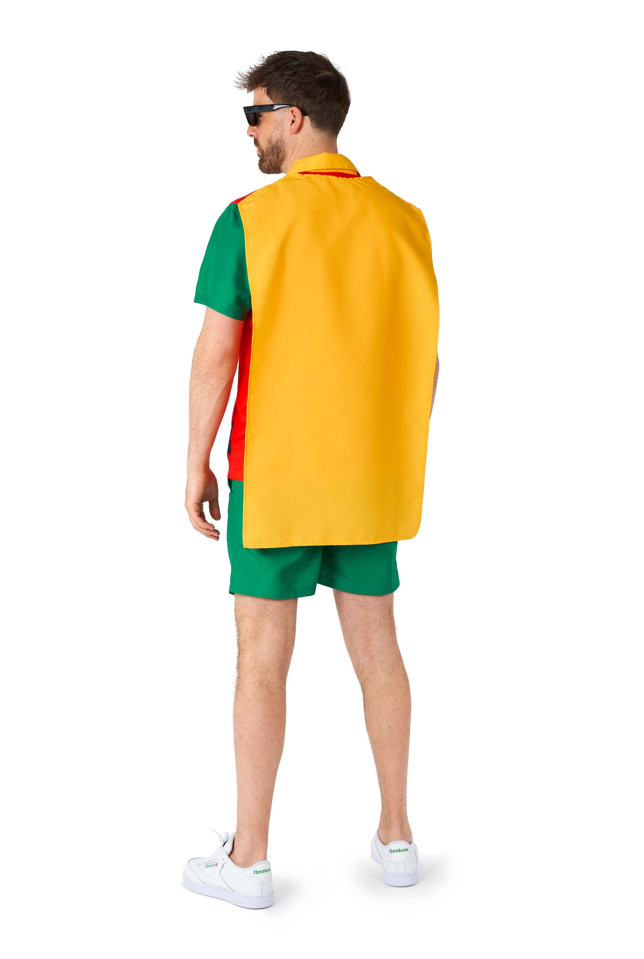 A man stands with his back turned, wearing a vibrant Robin costume from Suitmeister, featuring a striking yellow cape draped over his shoulders. The ensemble comprises a green shirt and shorts, complemented by white sneakers and sunglasses, giving him the appearance of a superhero.