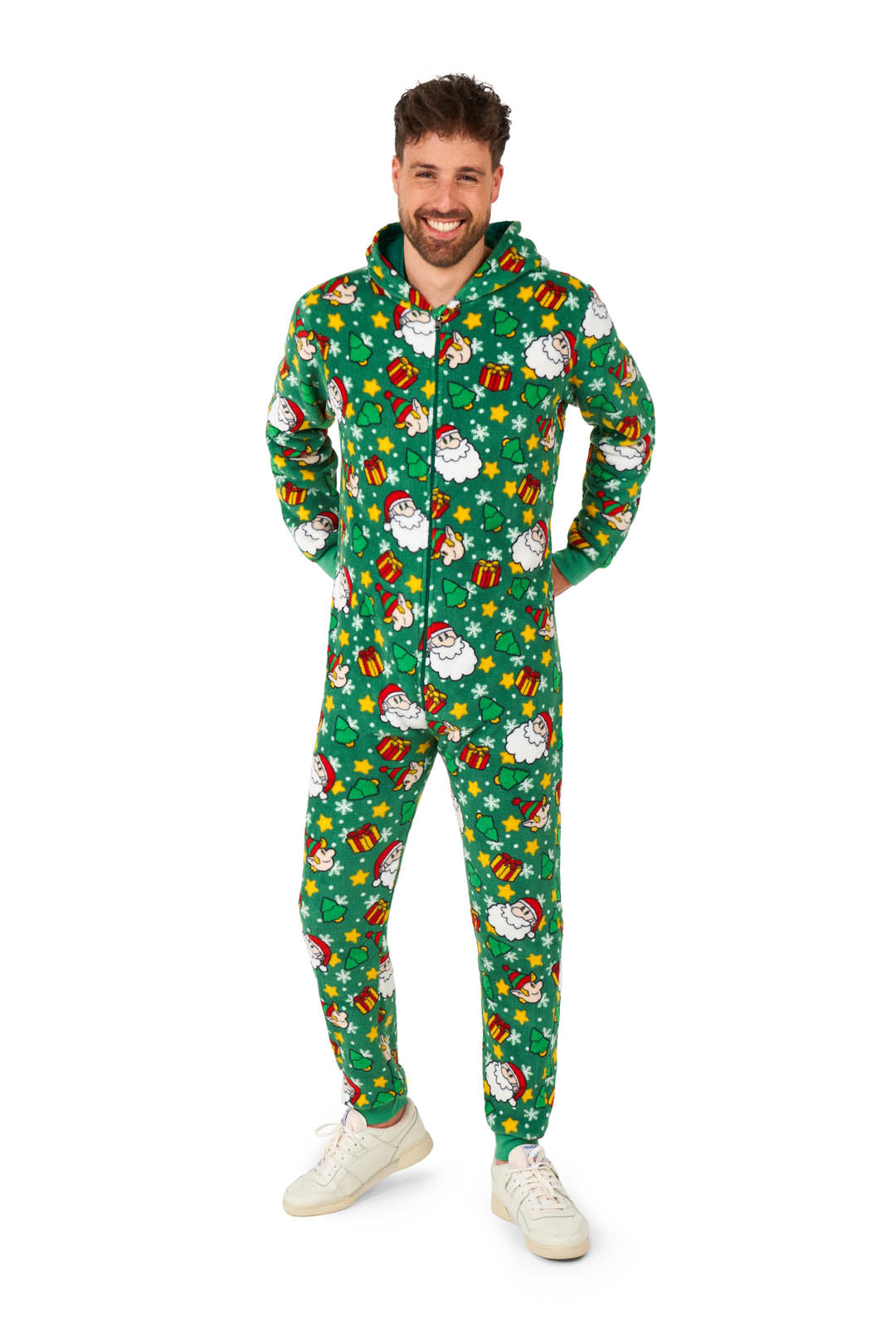 A person smiling and wearing a Suitmeister Santa Elves Green Christmas onesie featuring festive holiday patterns, such as snowmen and gifts. They have short hair and are standing against a white background with their hands in their pockets.