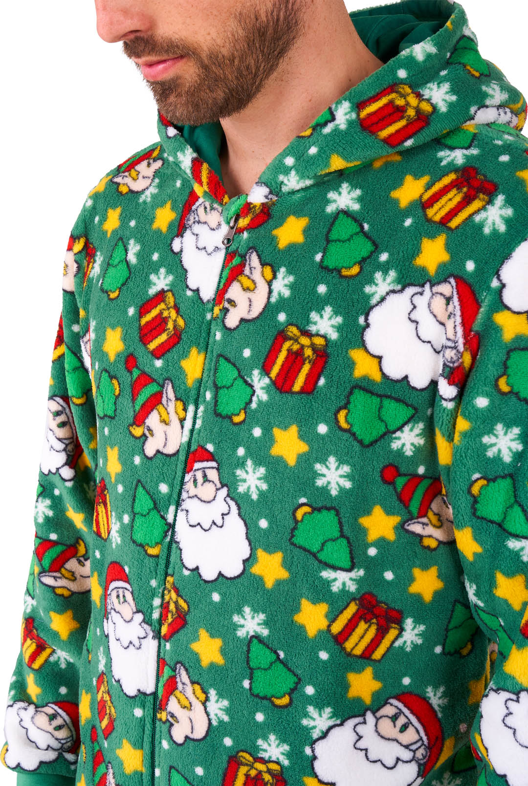 A person wearing the "Santa Elves Green - Christmas Onesie" by Suitmeister, featuring festive patterns such as stars, gift boxes, Santa faces, and Christmas trees. The onesie is primarily green with colorful accents, and the person's face is partially visible under the hood.