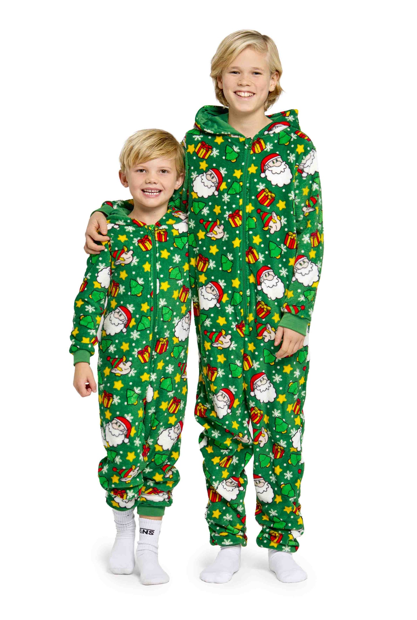 Two children wearing matching Santa Elves Green pajamas by Suitmeister stand side by side. The pajamas feature festive prints of Santa Claus, gifts, and snowflakes. The older child has an arm around the younger one, both smiling and wearing white socks.